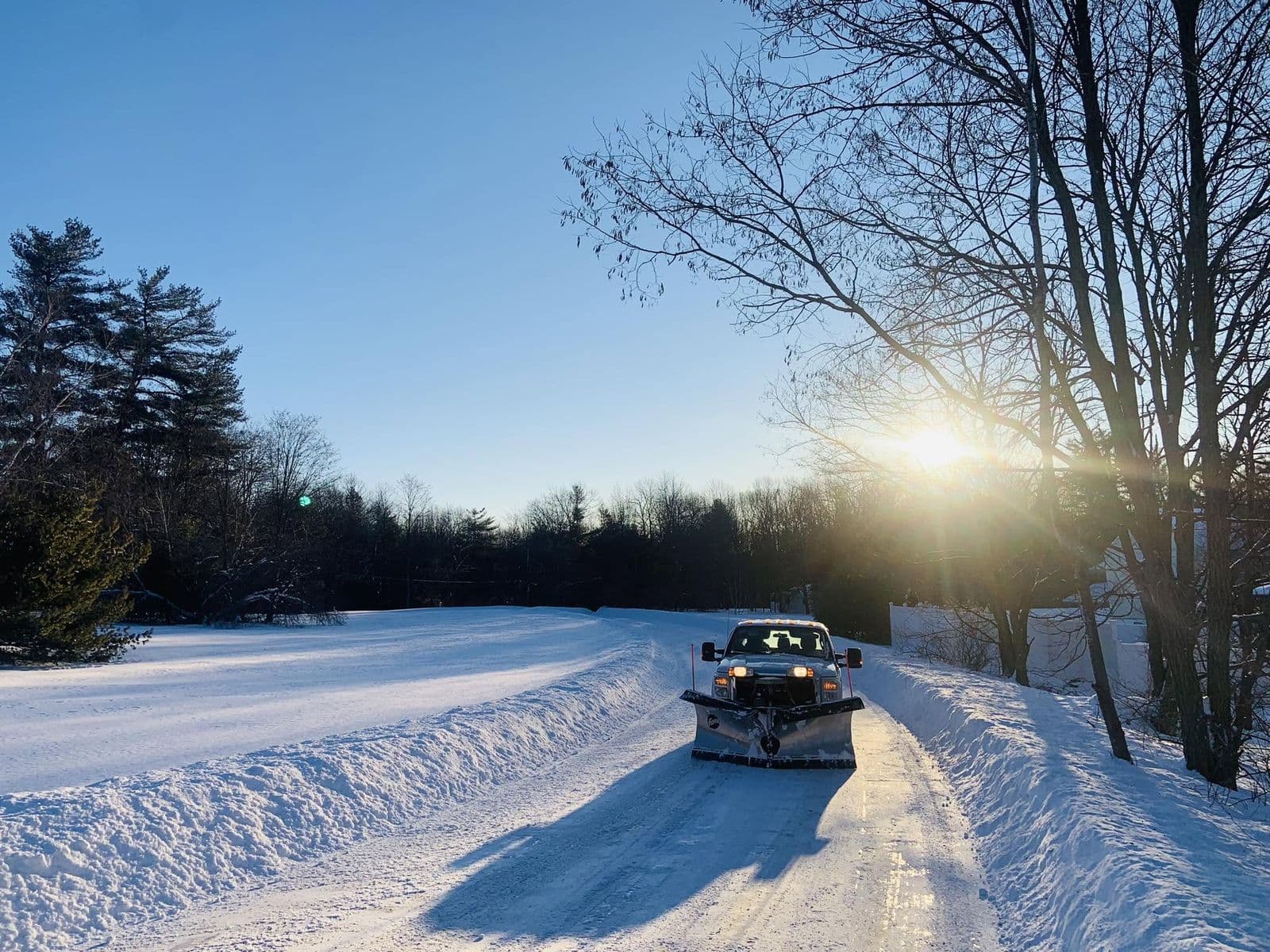 Pickup truck with a snowplow blade clearing a snow-covered rural driveway at sunrise